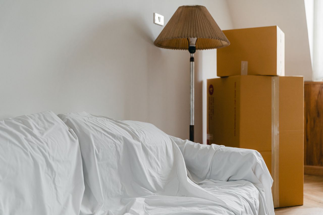 Sofa covered with a sheet, a floor lamp, and packed cardboard boxes in a bright, spacious room of a new contemporary house